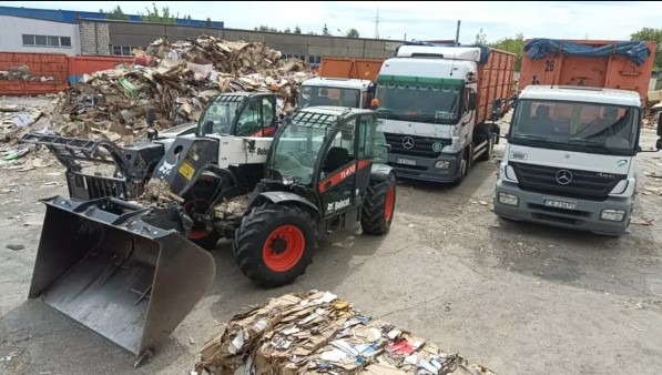 Dwa wózki widłowe Bobcat i dwie ciężarówki Mercedes na placu składowania makulatury w kostkach, widoczne kontenery i budynek przemysłowy w tle.