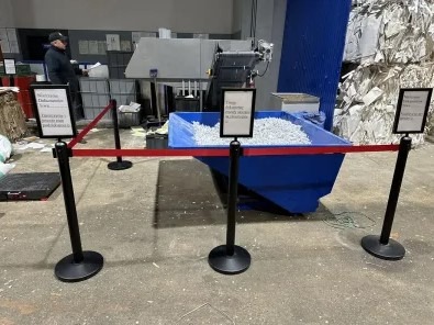 Linia odgradzająca z tabliczkami informacyjnymi przed niebieskim kontenerem wypełnionym pociętymi dokumentami, w tle maszyna do niszczenia dokumentów i sterta pociętych papierów.