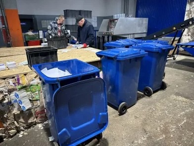 Utylizacja dokumentów: pracownicy sortują dokumenty nad stołem, obok niebieskie pojemniki na makulaturę, w tle taśmociąg.