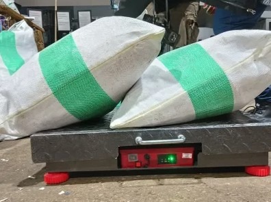 Dwa worki z zielonymi pasami umieszczone na wadze platformowej z czerwonym wyświetlaczem, w tle inne worki i fragmenty ubrań osób.