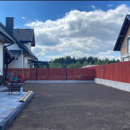 Równa trawka - Przygotowany teren pod trawnik między dwoma domami, widoczny niwelator laserowy na statywie, świeżo ułożony chodnik z kostki brukowej i drewniane deski w tle czerwone ogrodzenie.