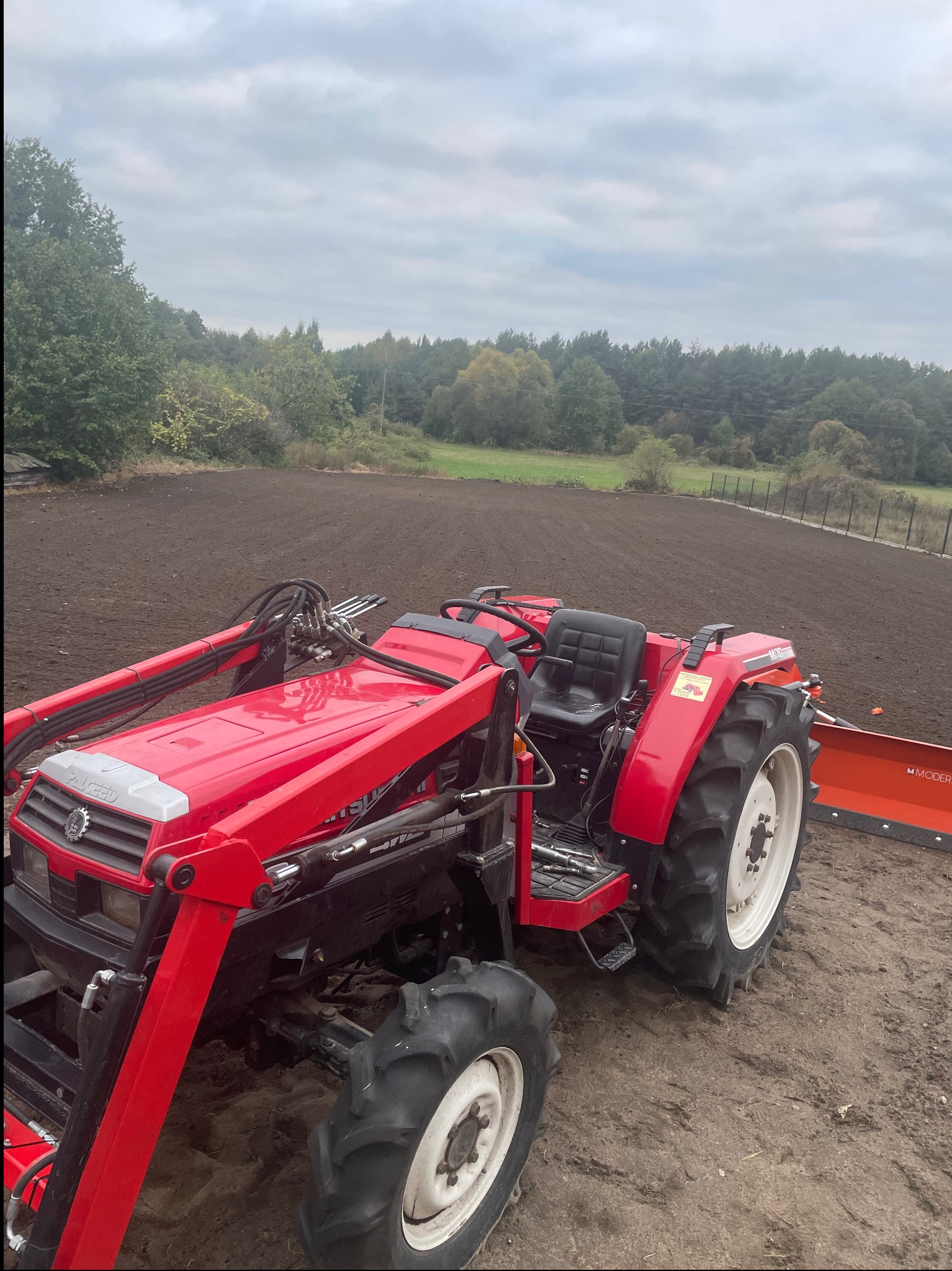 Czerwony traktor z przednim ładowaczem na świeżo zaoranej ziemi, przygotowanej pod uprawę, z lasem w tle i pochmurnym niebem.