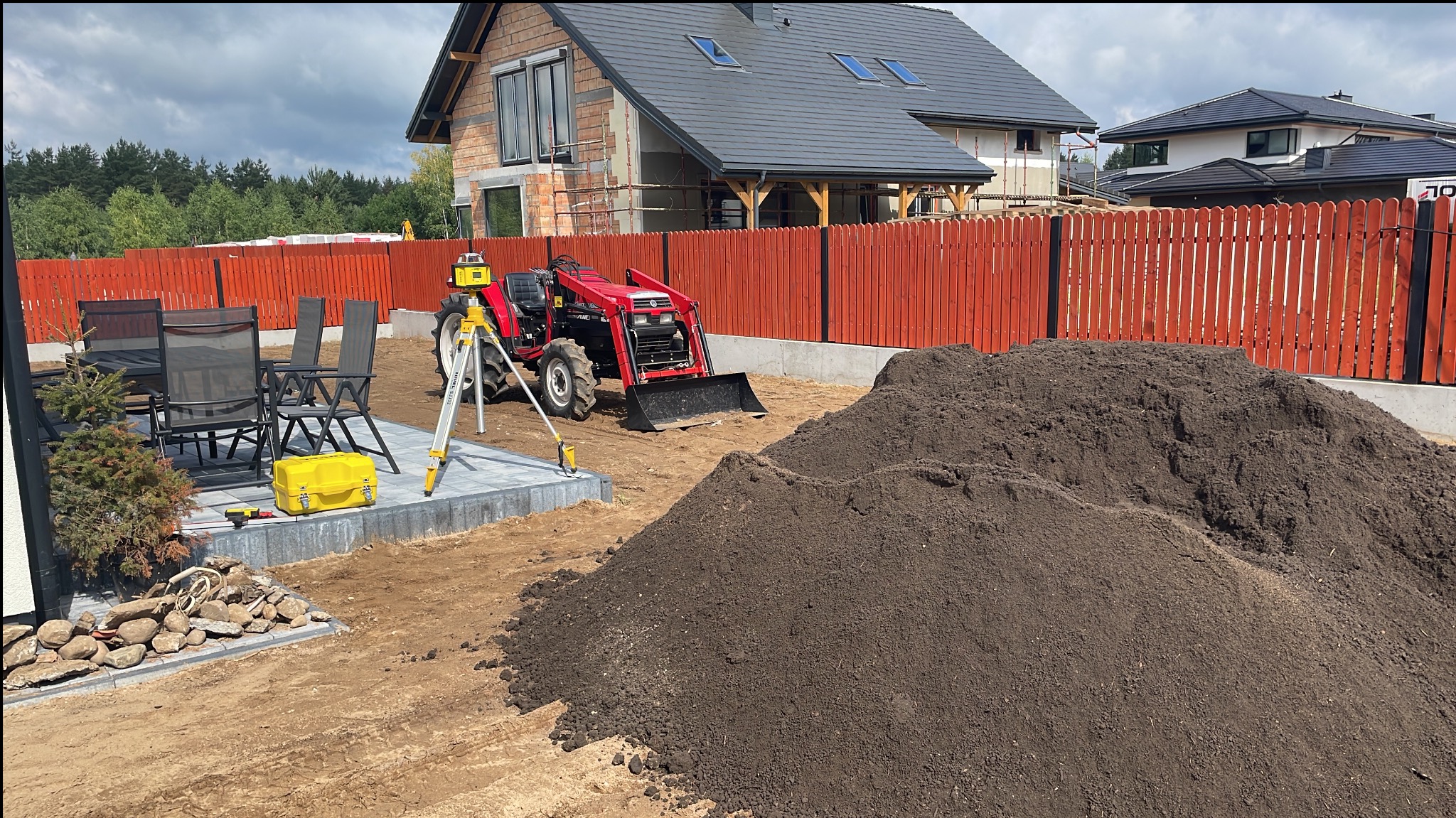 Czerwony traktor z łyżką stoi na wyrównanym terenie obok sterty ziemi, w tle częściowo wykończony dom z cegły i czerwone ogrodzenie, widoczny niwelator laserowy na statywie i zestaw mebli...
