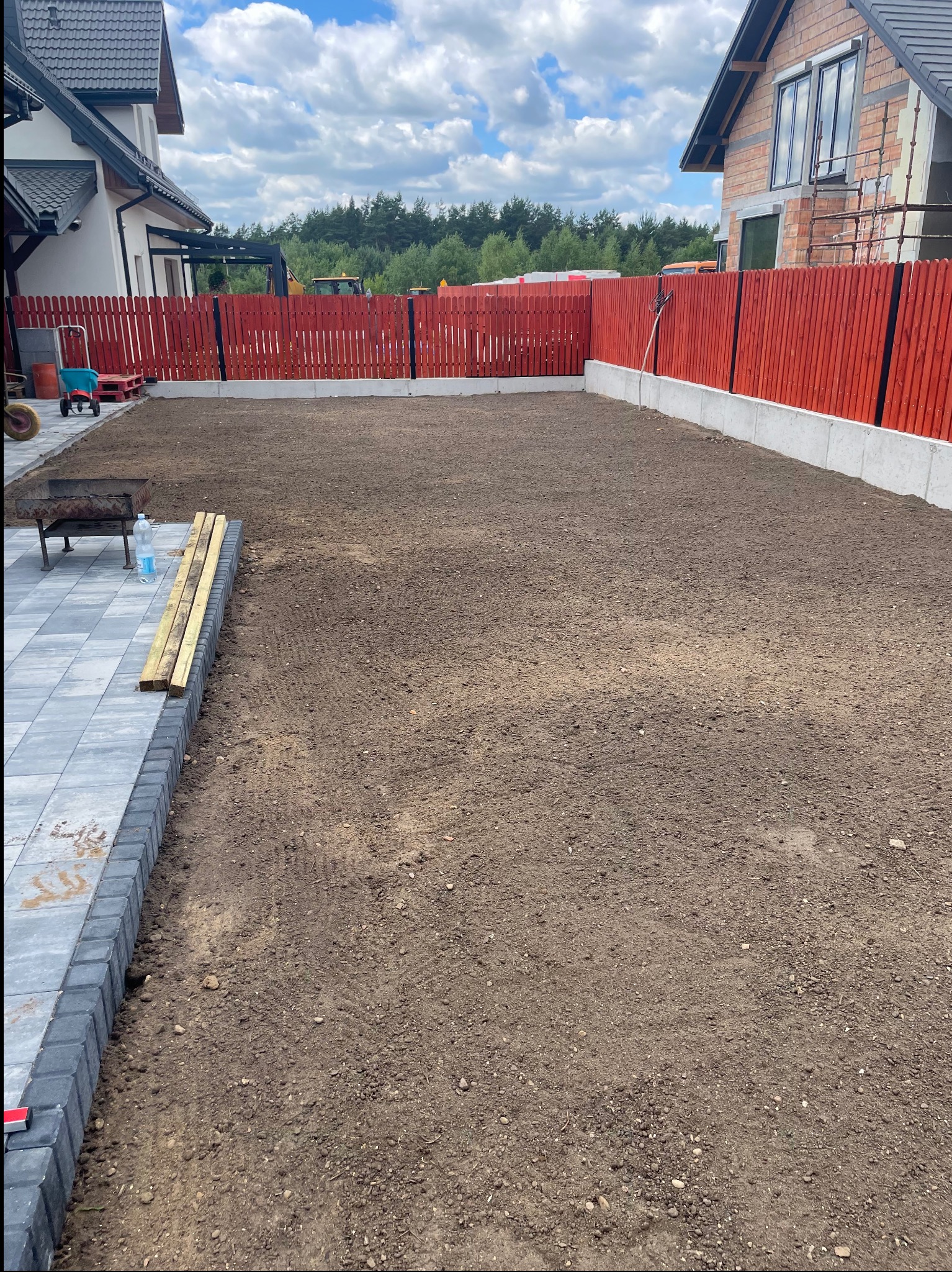 Przygotowany teren pod przyszły trawnik, widoczna świeżo wyrównana ziemia, obrzeża z kostki brukowej, czerwone ogrodzenie i budynki w tle. W oddali widoczny sprzęt budowlany.