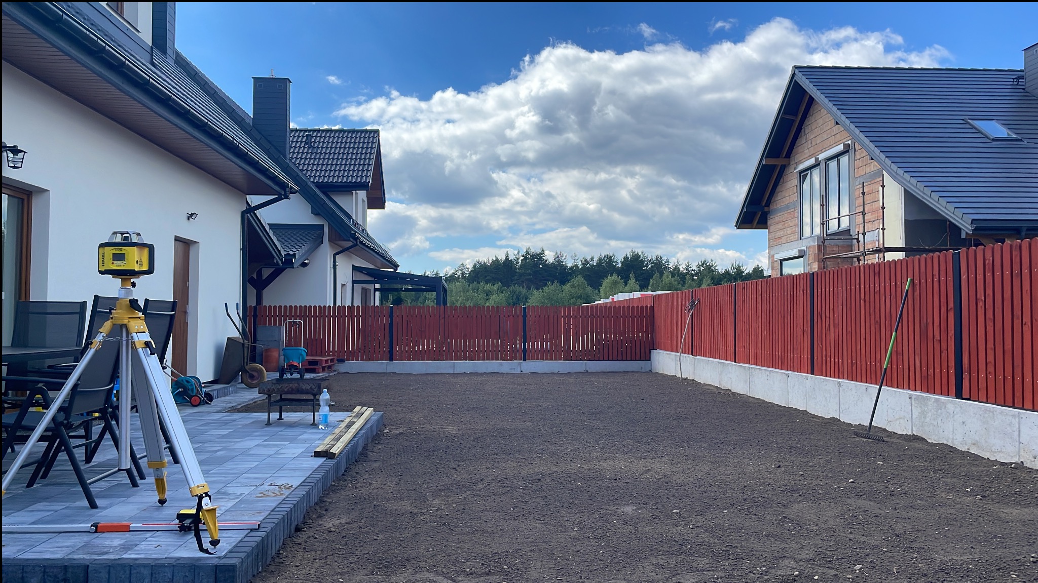 Przygotowany teren pod trawnik między dwoma domami, widoczny niwelator laserowy na statywie, świeżo ułożony chodnik z kostki brukowej i drewniane deski w tle czerwone ogrodzenie.