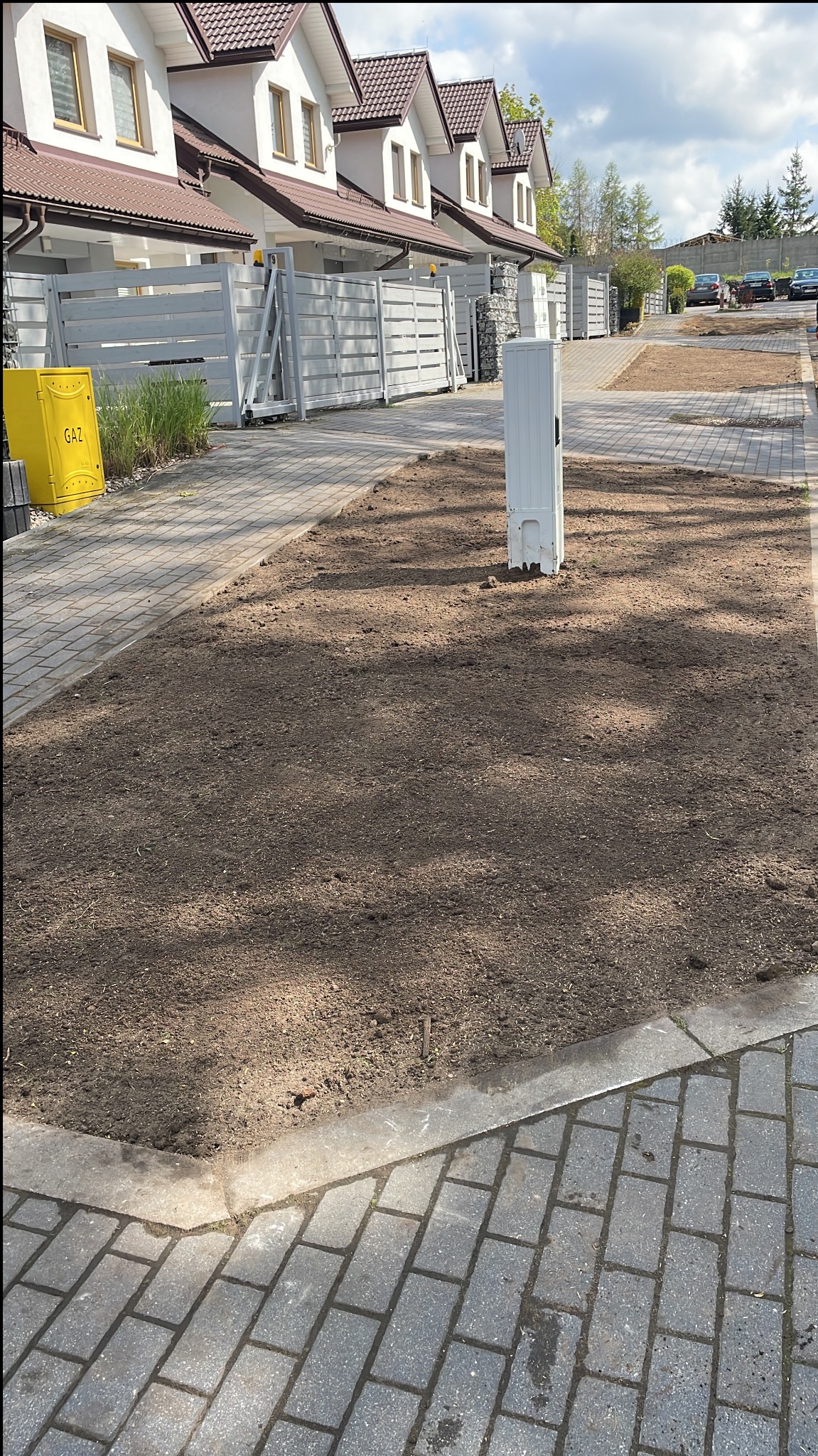 Przygotowany teren pod przyszły trawnik, świeżo wyrównana ziemia pomiędzy chodnikiem z kostki brukowej a zabudową szeregową.