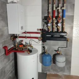 Nowoczesna kotłownia z białym piecem kondensacyjnym, zasobnikiem ciepłej wody, rozdzielaczem z pompami obiegowymi i naczyniami wzbiorczymi, na tle ściany wyłożonej płytkami imitującymi marmur.