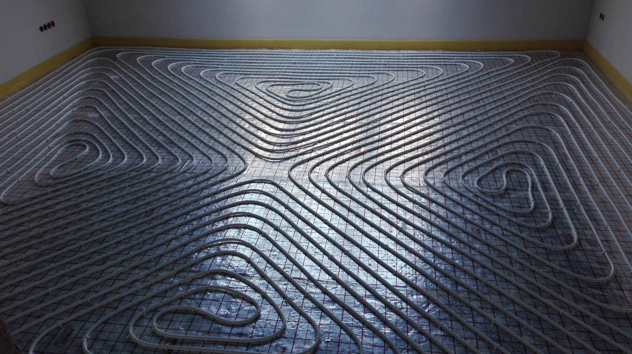 Białe rury ogrzewania podłogowego ułożone w spiralny wzór na folii izolacyjnej z siatką, widoczne w nowym pomieszczeniu z żółtą taśmą przy ścianach.