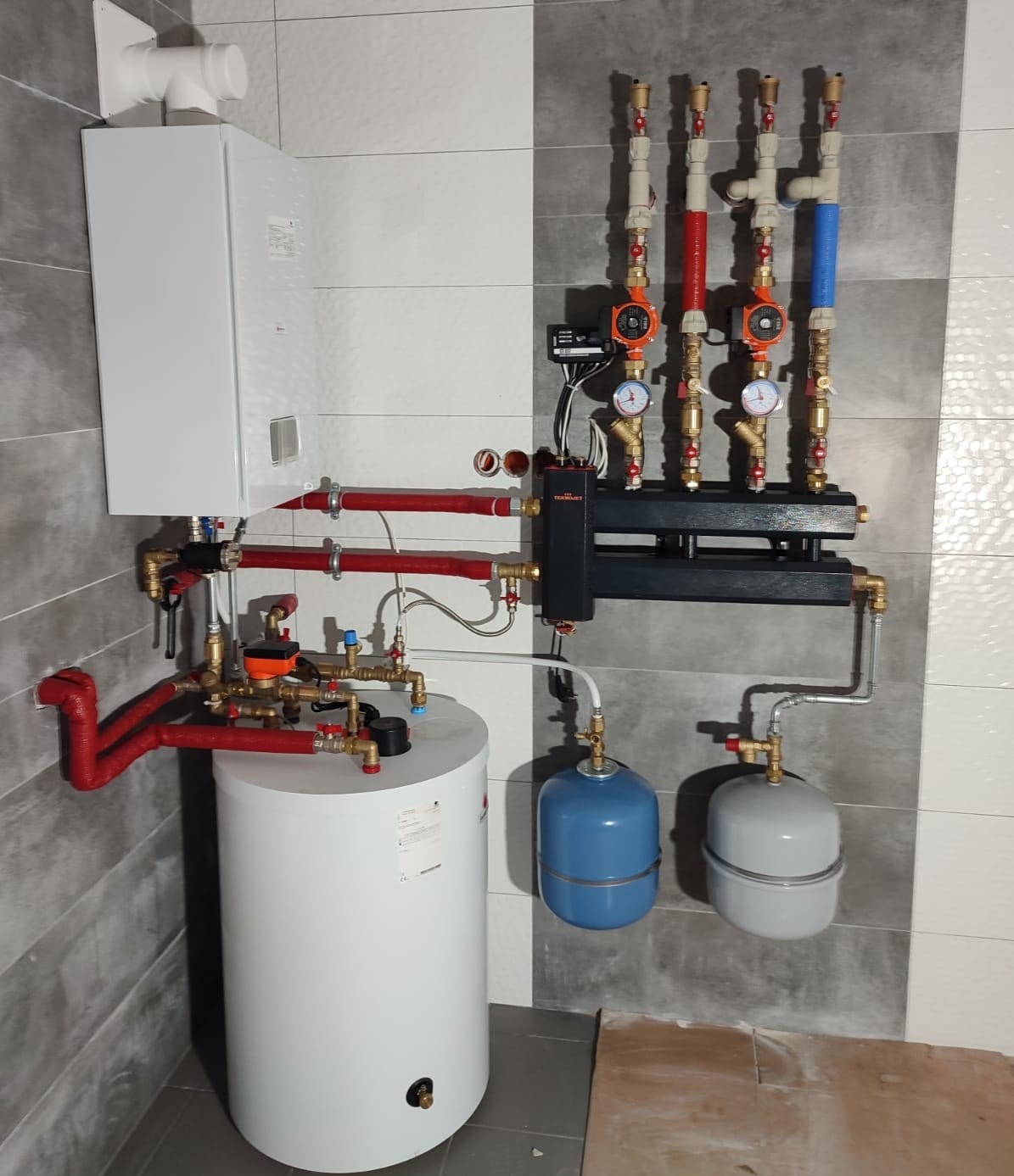 Nowoczesna kotłownia z białym piecem kondensacyjnym, zasobnikiem ciepłej wody, rozdzielaczem z pompami obiegowymi i naczyniami wzbiorczymi, na tle ściany wyłożonej płytkami imitującymi marmur.