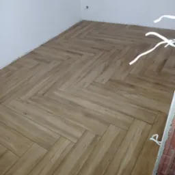 Ułożona pod kątem jodełki jasnobrązowa podłoga z paneli imitujących drewno, widoczne fragmenty białej ściany i okna zabezpieczonego folią malarską.