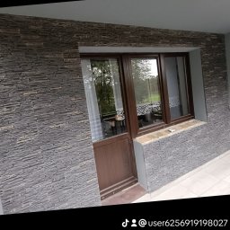 Oleksander Gryszczuk - Elewacja budynku z kamienną okładziną, brązowe okno z widokiem na zieleń, biała firanka.