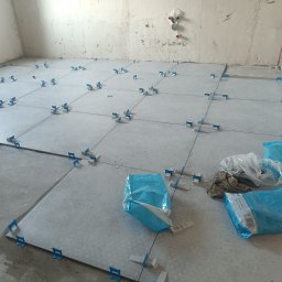 Oleksander Gryszczuk - W trakcie prac: Układanie jasnoszarych płytek podłogowych z użyciem niebieskich klipsów poziomujących, widoczne worki z klejem i nieprzygotowane ściany.