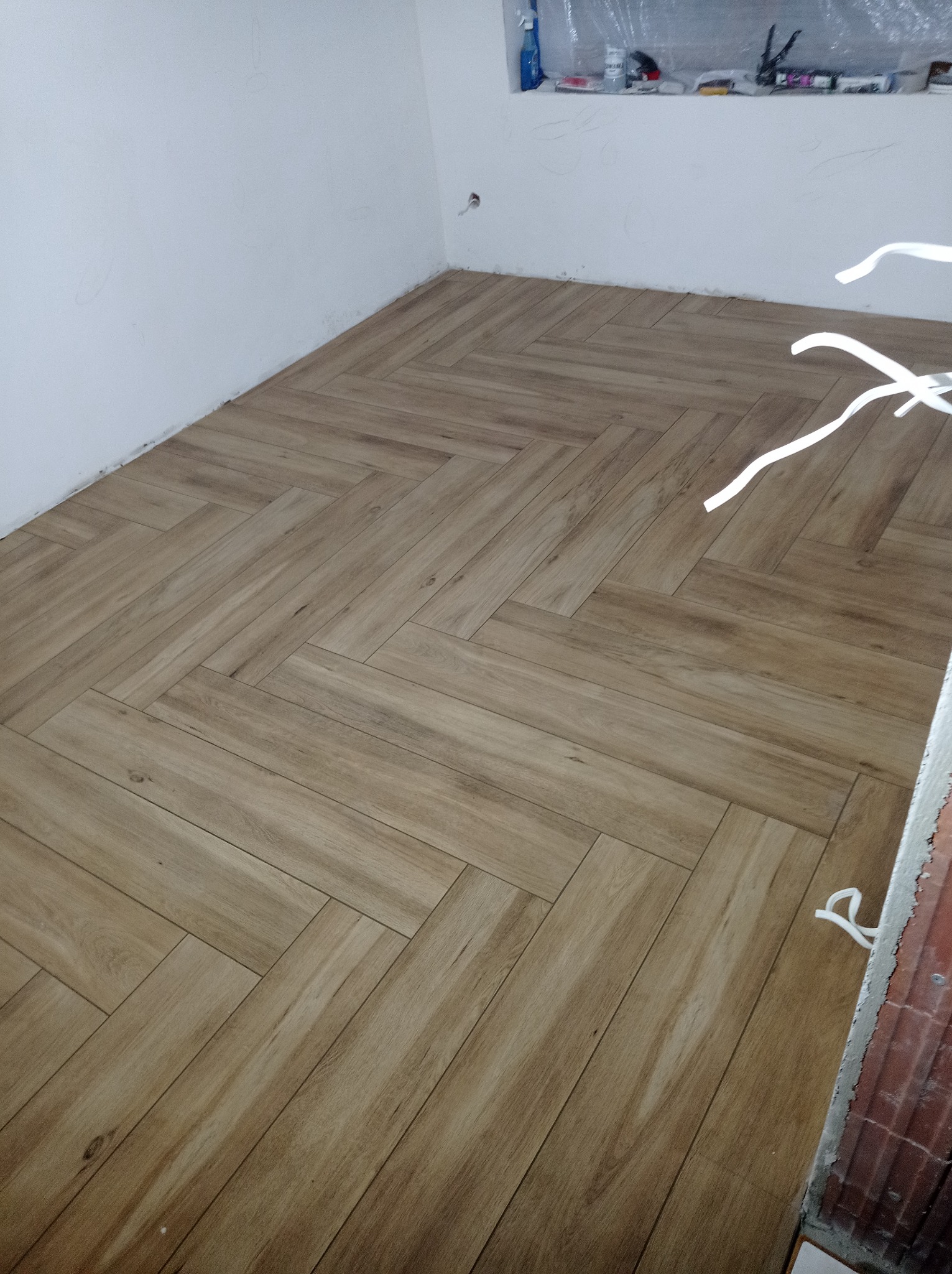 Ułożona pod kątem jodełki jasnobrązowa podłoga z paneli imitujących drewno, widoczne fragmenty białej ściany i okna zabezpieczonego folią malarską.