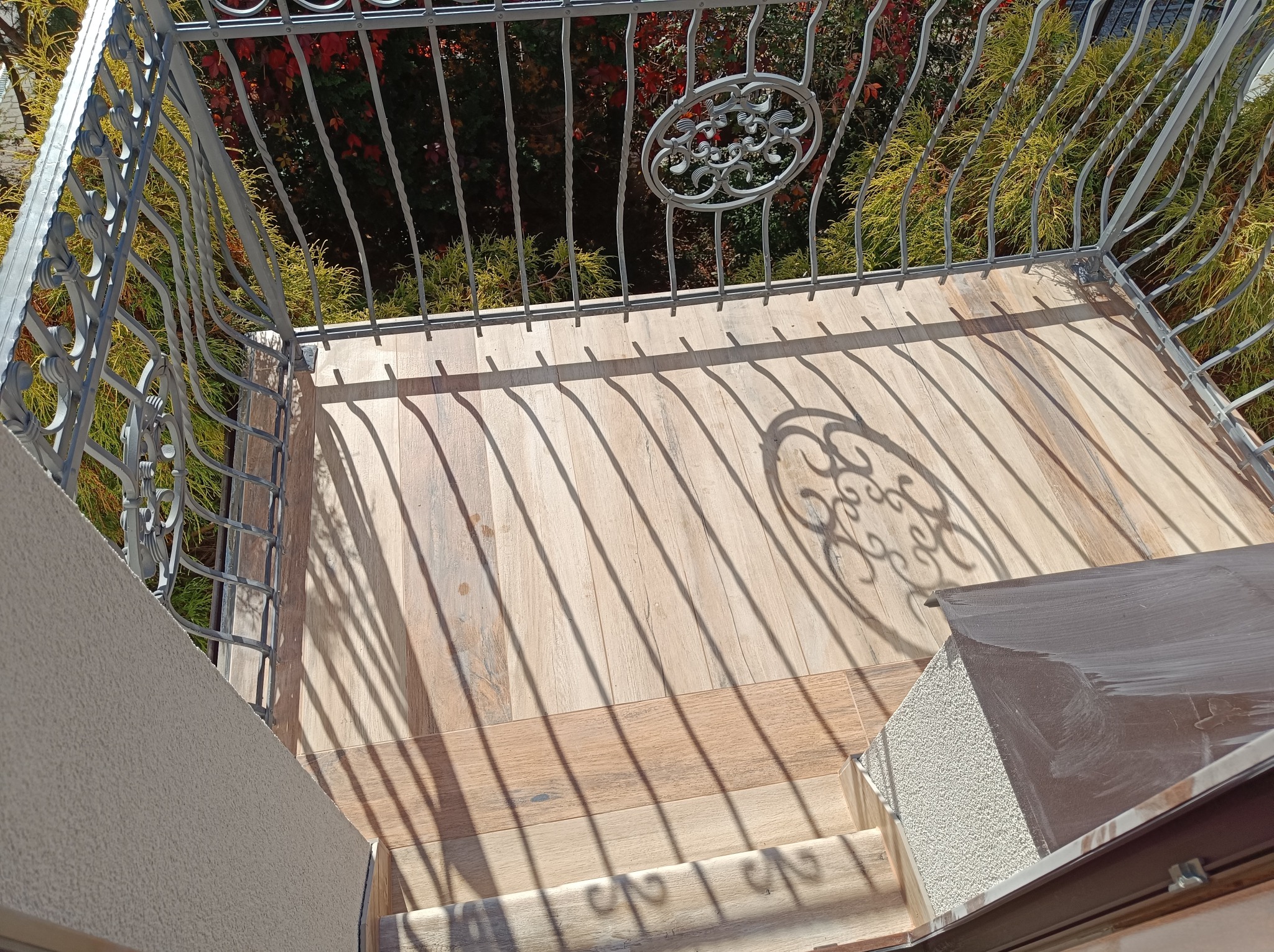 Balkon wyłożony płytkami imitującymi drewno, z metalową balustradą rzucającą cienie na podłogę.