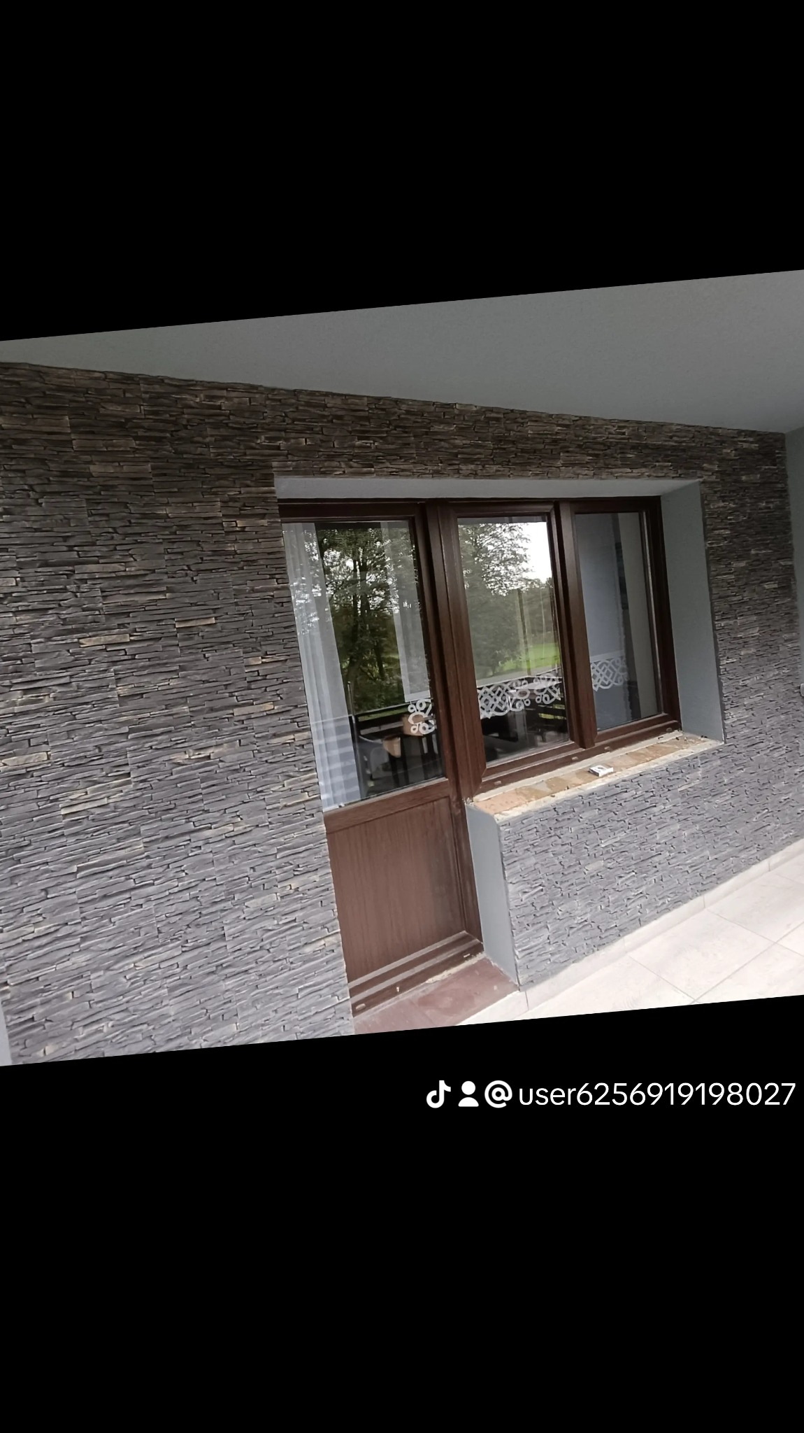 Elewacja budynku z kamienną okładziną, brązowe okno z widokiem na zieleń, biała firanka.