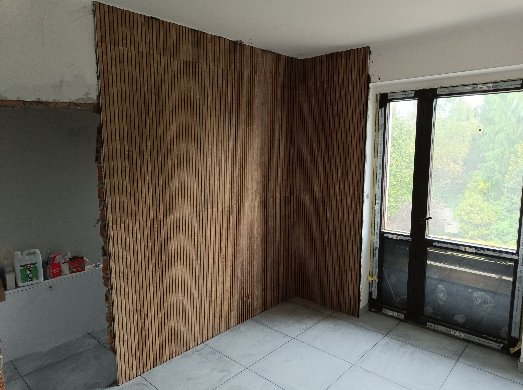 Montaż drewnianych paneli ściennych w pomieszczeniu z oknem, widoczne niedokończone prace przy ścianie obok wejścia, szare płytki podłogowe.