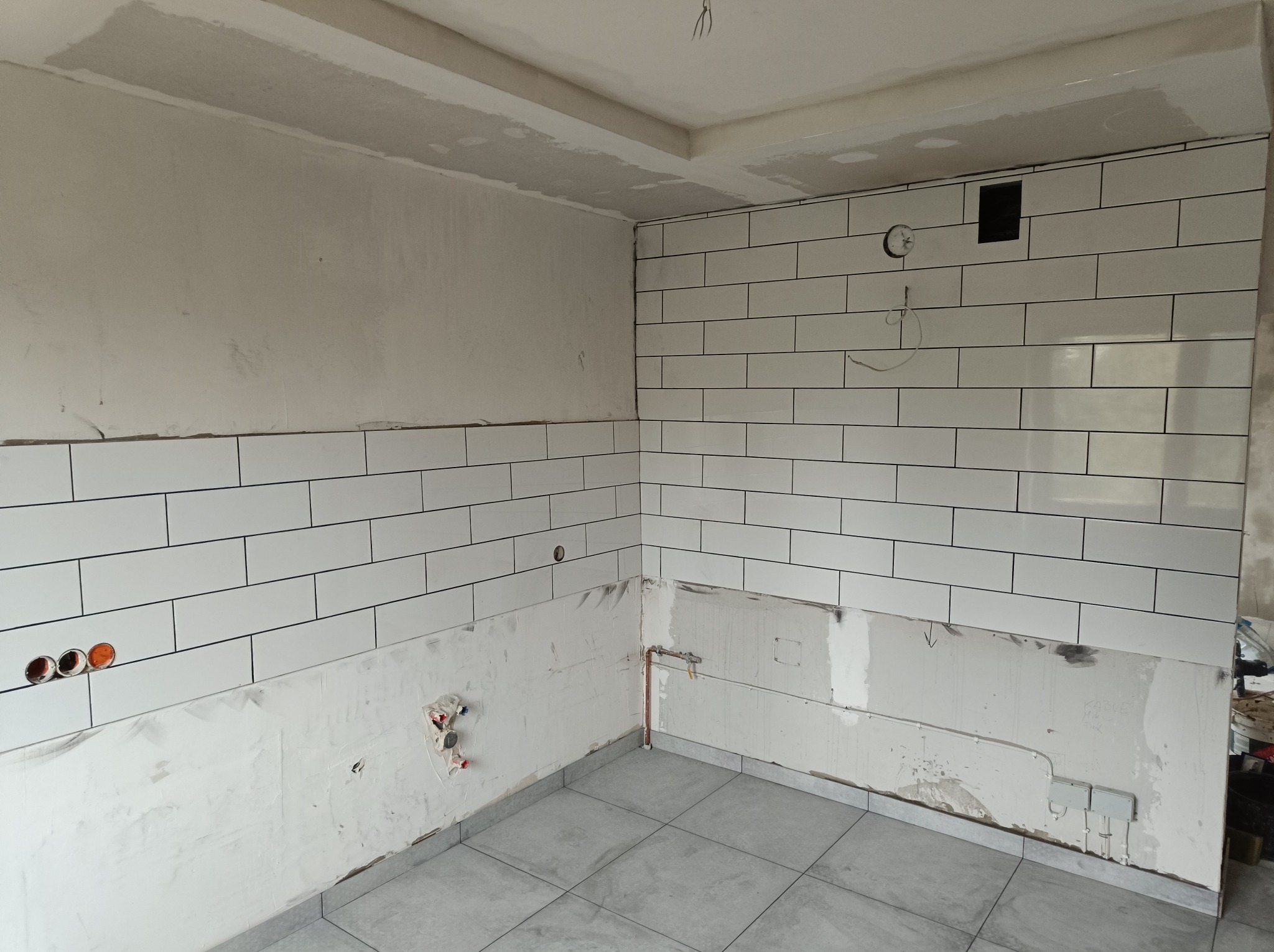 Wnętrze łazienki w Cieszynie w trakcie prac: białe płytki ułożone do połowy ściany, widoczne instalacje elektryczne i hydrauliczne, surowy stan wykończenia.