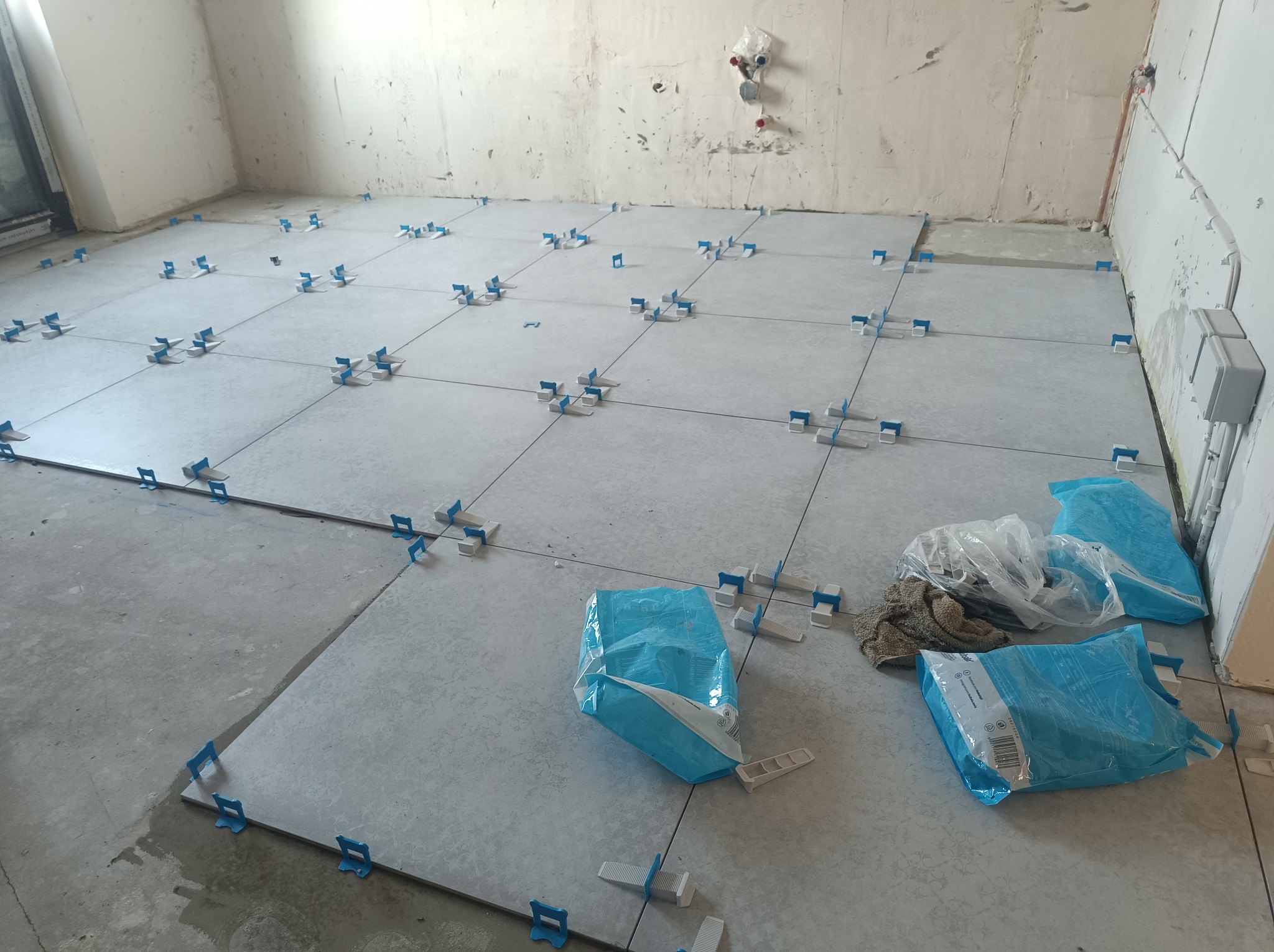 W trakcie prac: Układanie jasnoszarych płytek podłogowych z użyciem niebieskich klipsów poziomujących, widoczne worki z klejem i nieprzygotowane ściany.