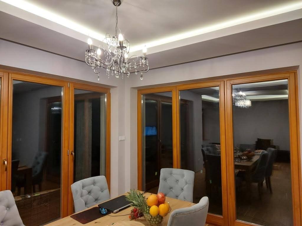 Jasne wnętrze jadalni z drewnianymi oknami, kryształowym żyrandolem i stołem z owocami, widoczne detale wykończenia i oświetlenie sufitowe.