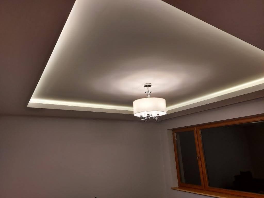 Nowoczesny sufit podwieszany z oświetleniem LED, centralnie umieszczony żyrandol z abażurem, widoczne okno z drewnianą ramą.