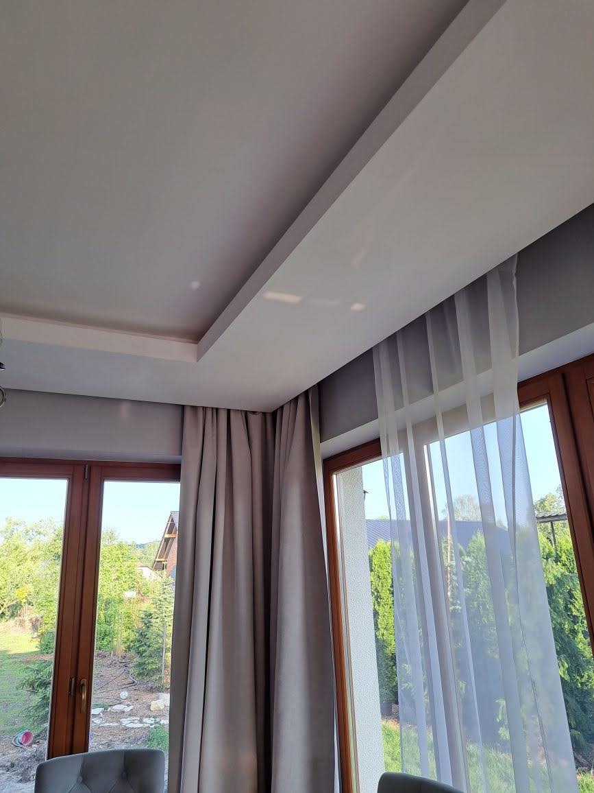 Nowoczesny sufit podwieszany z listwą oświetleniową i zasłonami w odcieniach szarości, widok na ogród przez drewniane okna.