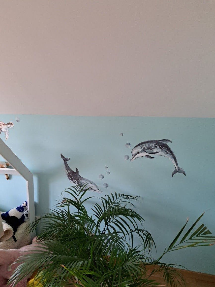Dwa delfiny jako naklejki na błękitnej ścianie w pokoju dziecięcym, z zieloną rośliną doniczkową na pierwszym planie i fragmentem białej ramy łóżka z zabawkami w tle.