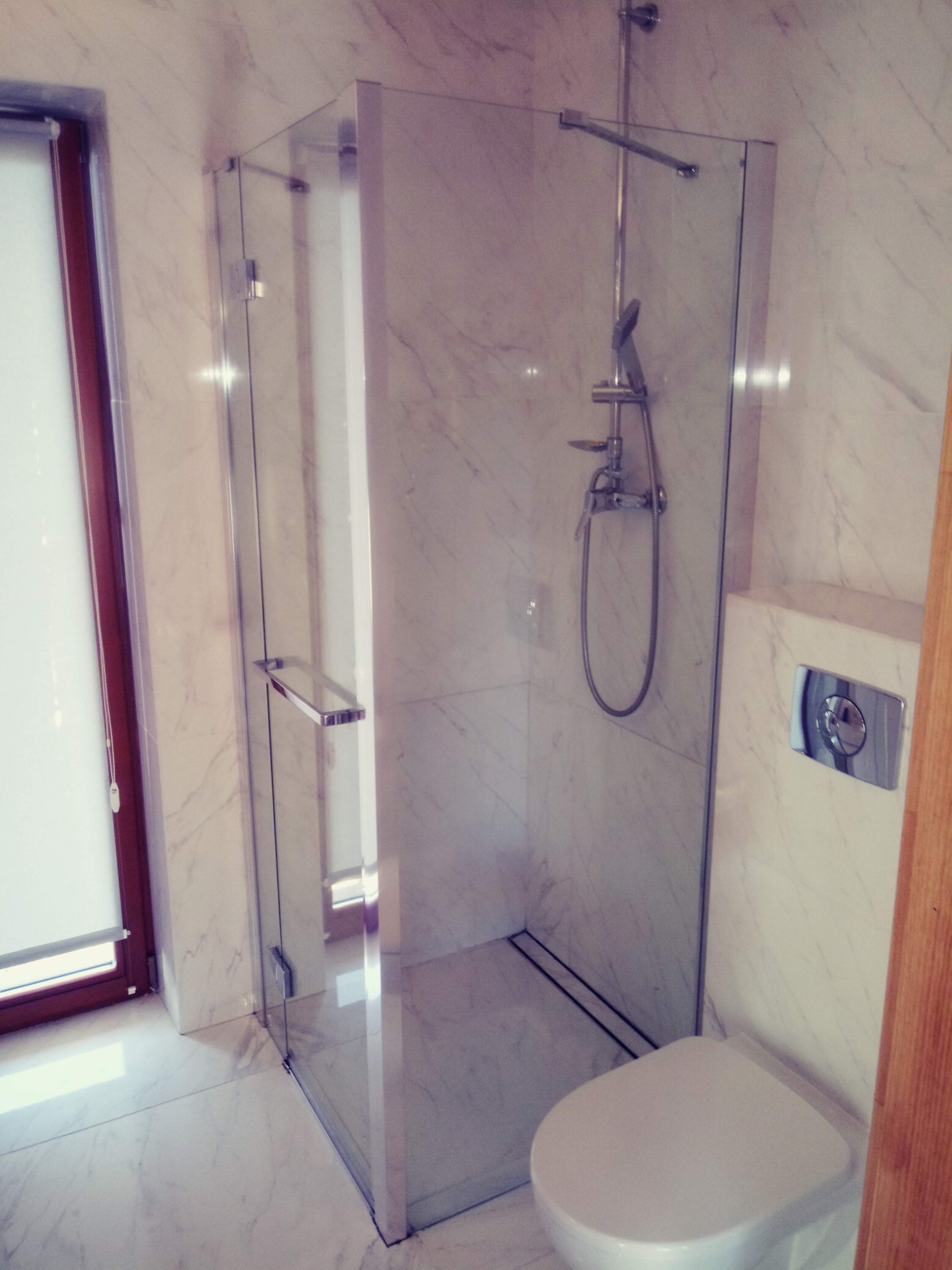 Nowoczesna kabina prysznicowa z transparentnego szkła w łazience wykończonej białym marmurem, widoczny chromowany zestaw prysznicowy i wisząca toaleta z przyciskiem spłukującym w kolorze srebrnym.