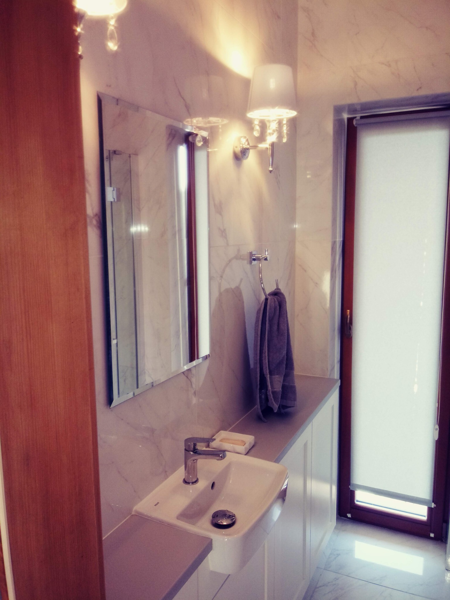 Nowoczesna łazienka w Tenczynku z marmurowymi płytkami, wiszącą umywalką z chromowaną baterią, lustrem w srebrnej ramie i dekoracyjnym oświetleniem ściennym.