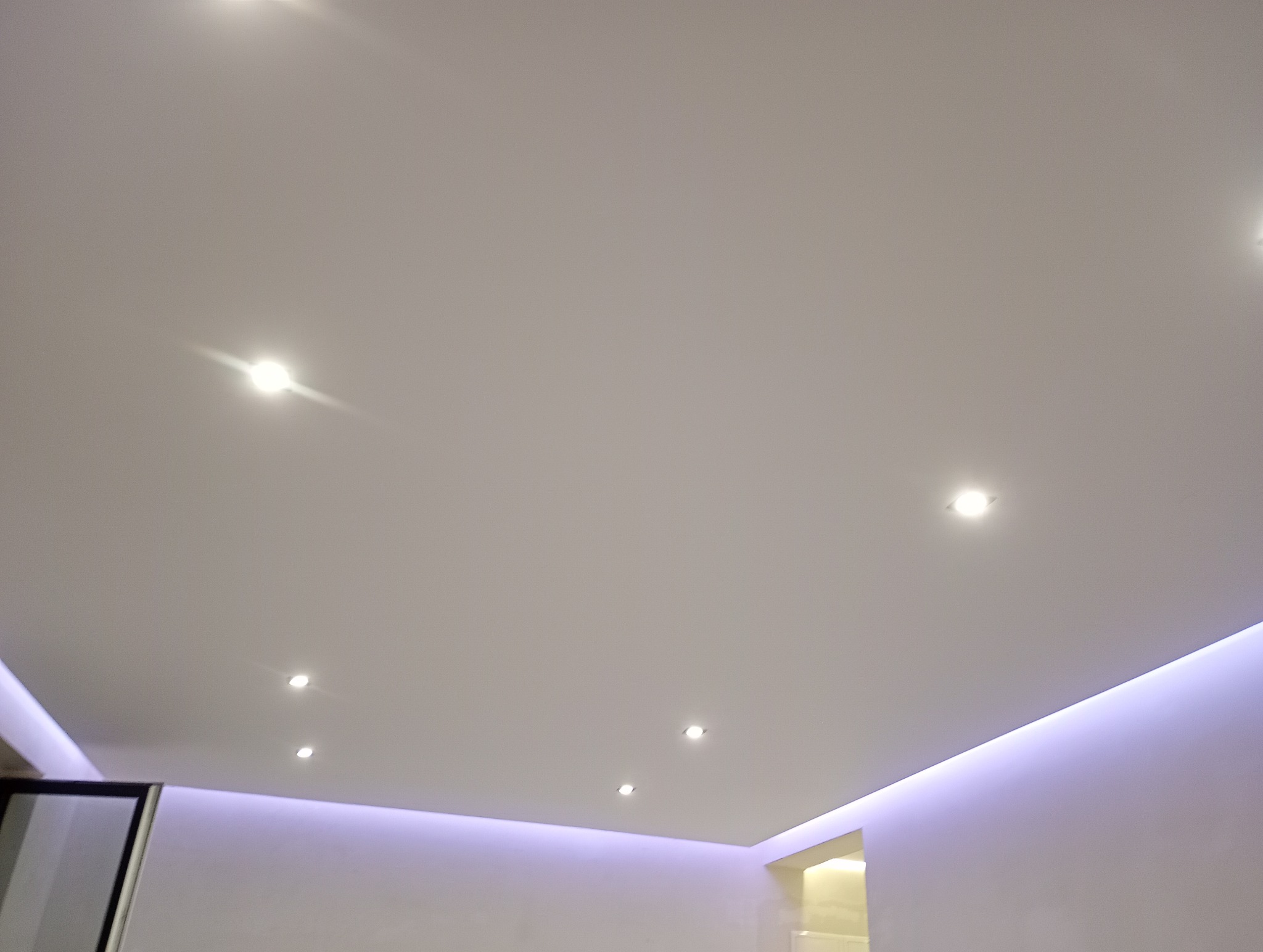 Gładki, biały sufit z ukrytymi listwami LED emitującymi fioletowe światło i wpuszczonymi punktowymi lampami o ciepłej barwie.