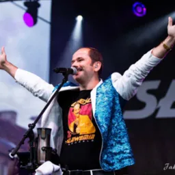 Mężczyzna z wąsami w koszulce z wizerunkiem Sławomira, w białej marynarce z niebieskimi cekinami, śpiewa do mikrofonu na scenie z uniesionymi rękami, oświetlony reflektorami.