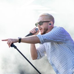 Jakub Wieczorek - Wokalista z blond włosami w okularach przeciwsłonecznych, śpiewa do mikrofonu, wskazując palcem w trakcie występu na scenie.