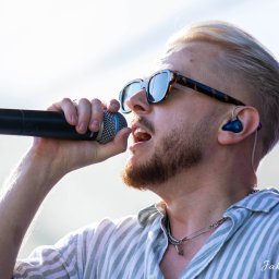 Jakub Wieczorek - Młody mężczyzna z blond włosami i brodą, w okularach przeciwsłonecznych, śpiewa do mikrofonu, nosząc koszulę w paski i słuchawkę douszną.