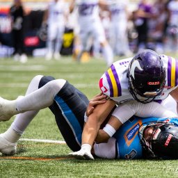 Jakub Wieczorek - Starcie zawodników futbolu amerykańskiego podczas meczu w Legnicy: jeden zawodnik w fioletowo-białym stroju powala drugiego w niebieskim stroju na zieloną murawę, w tle rozmyte sylwetki innych graczy.
