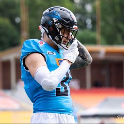 Jakub Wieczorek - Zawodnik futbolu amerykańskiego w niebieskiej koszulce i czarnym kasku z wizerunkiem smoka, poprawiający rękawicę na tle stadionu.