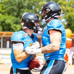 Jakub Wieczorek - Dwóch zawodników futbolu amerykańskiego w jasnoniebieskich strojach i kaskach, jeden z tatuażem na ramieniu, trzyma piłkę do futbolu amerykańskiego.