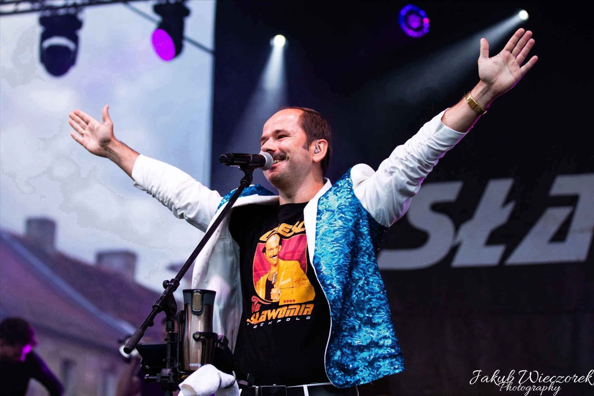 Mężczyzna z wąsami w koszulce z wizerunkiem Sławomira, w białej marynarce z niebieskimi cekinami, śpiewa do mikrofonu na scenie z uniesionymi rękami, oświetlony reflektorami.