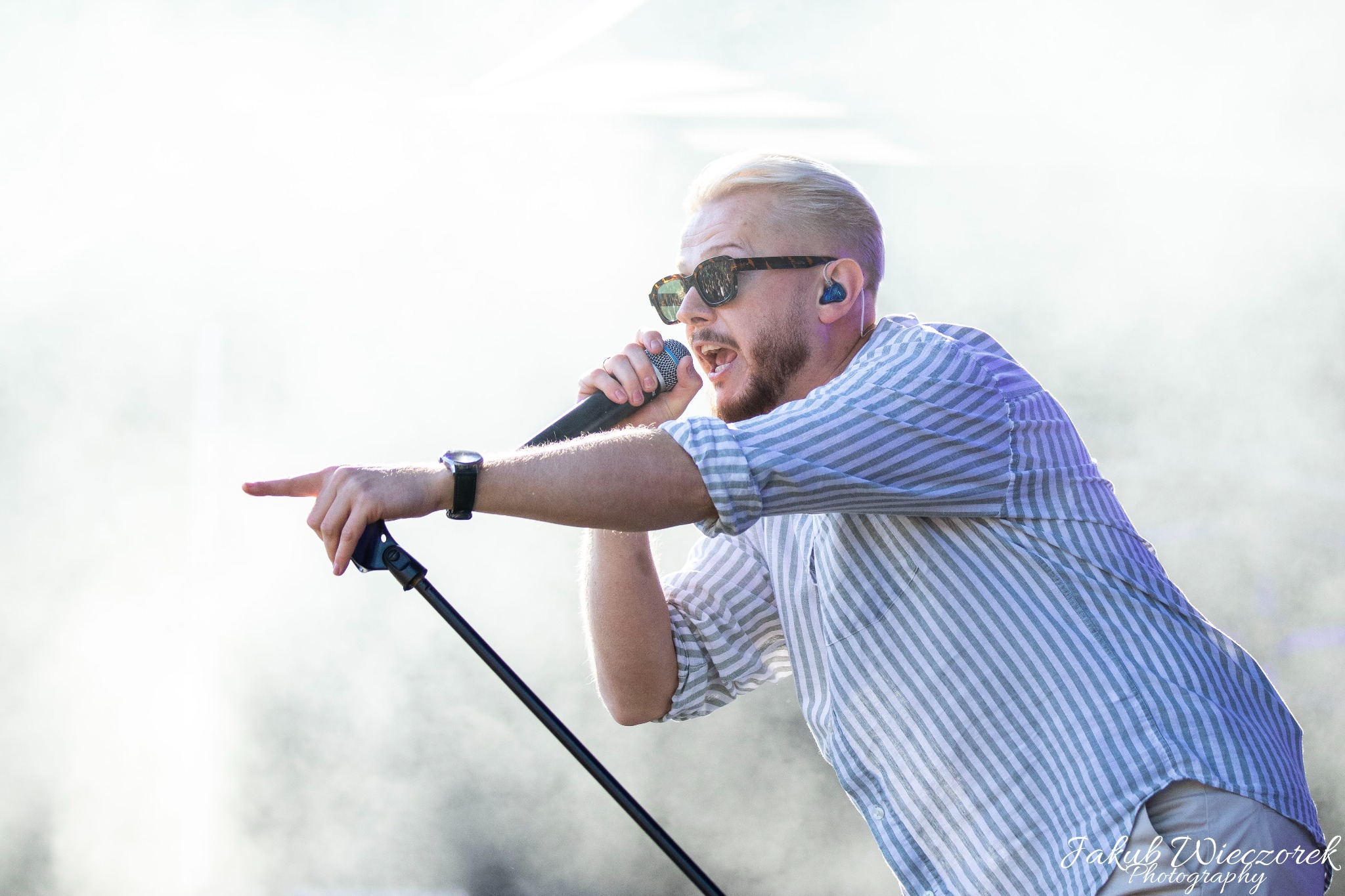 Wokalista z blond włosami w okularach przeciwsłonecznych, śpiewa do mikrofonu, wskazując palcem w trakcie występu na scenie.