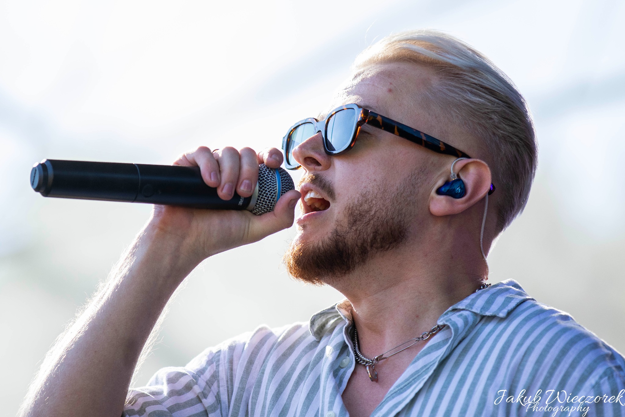 Młody mężczyzna z blond włosami i brodą, w okularach przeciwsłonecznych, śpiewa do mikrofonu, nosząc koszulę w paski i słuchawkę douszną.