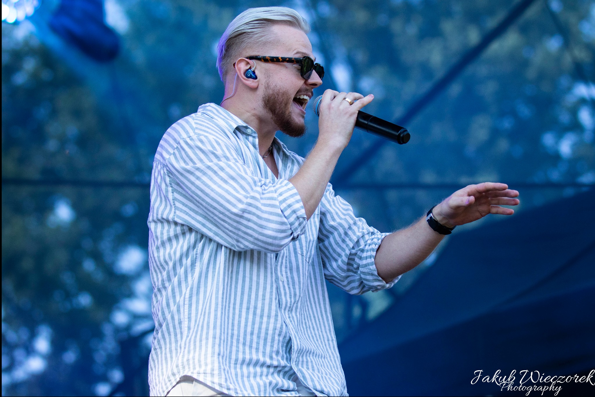 Wokalista z blond włosami i brodą, w okularach przeciwsłonecznych, śpiewa do mikrofonu na scenie podczas eventu, ubrany w koszulę w paski.