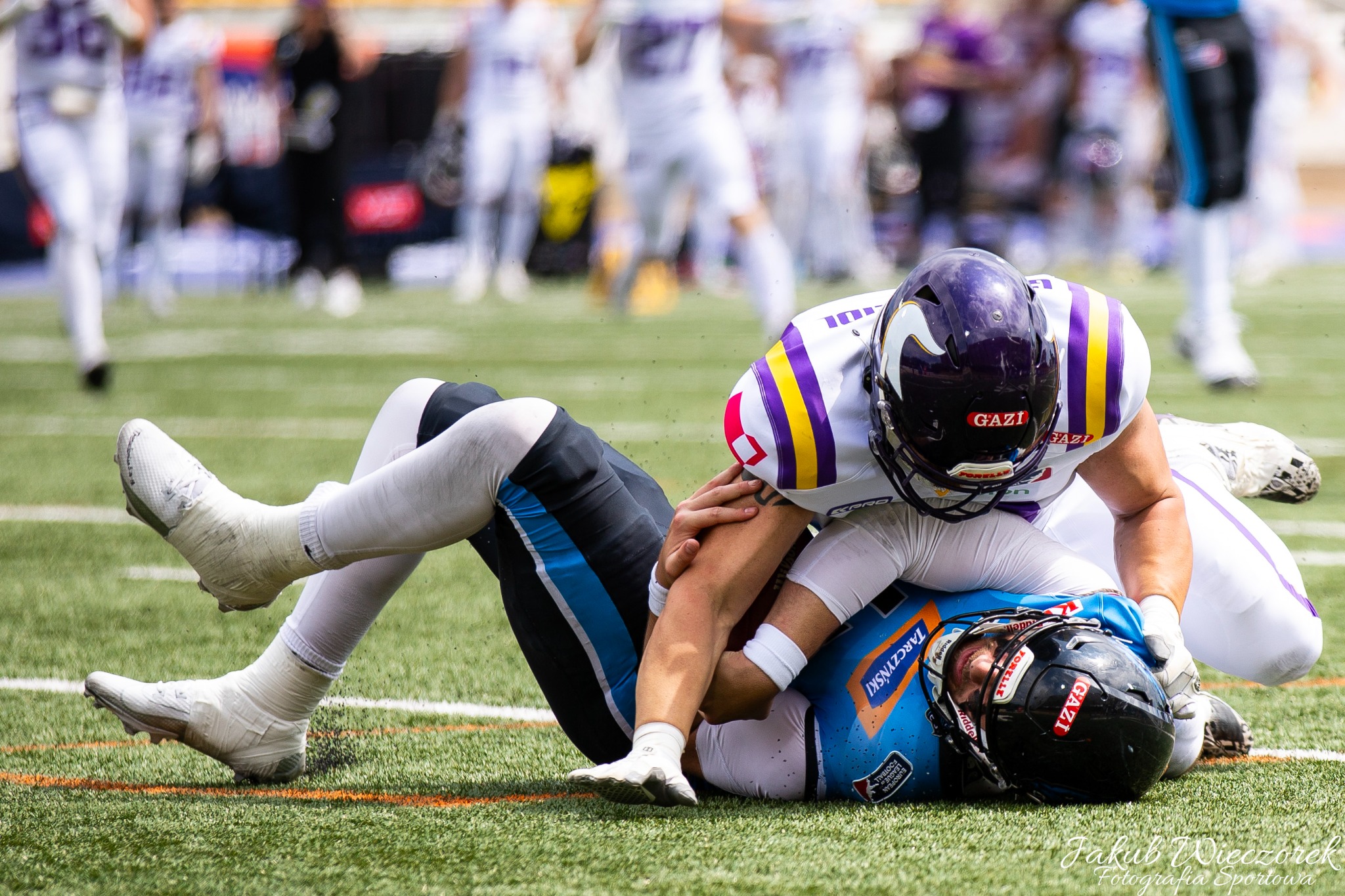 Starcie zawodników futbolu amerykańskiego podczas meczu w Legnicy: jeden zawodnik w fioletowo-białym stroju powala drugiego w niebieskim stroju na zieloną murawę, w tle rozmyte sylwetki innych graczy.