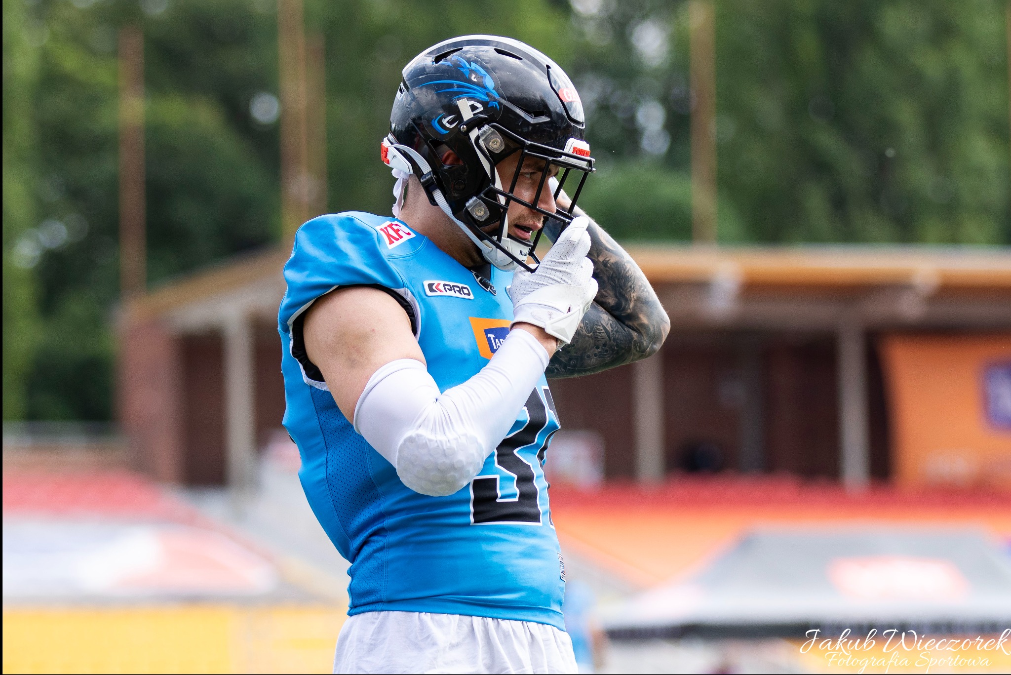 Zawodnik futbolu amerykańskiego w niebieskiej koszulce i czarnym kasku z wizerunkiem smoka, poprawiający rękawicę na tle stadionu.