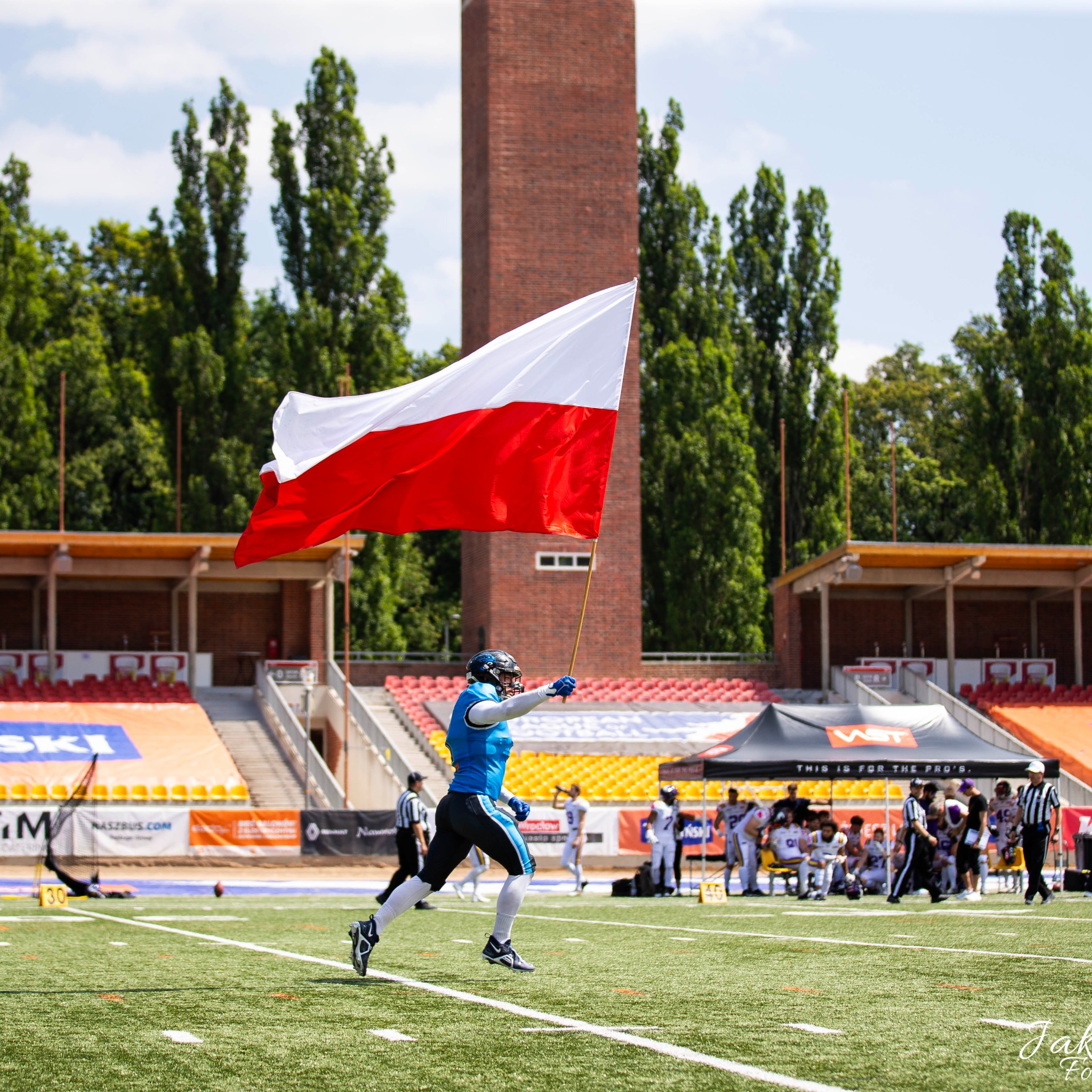 Zawodnik futbolu amerykańskiego biegnie po boisku z polską flagą na kiju, na tle trybun i wieży ciśnień.
