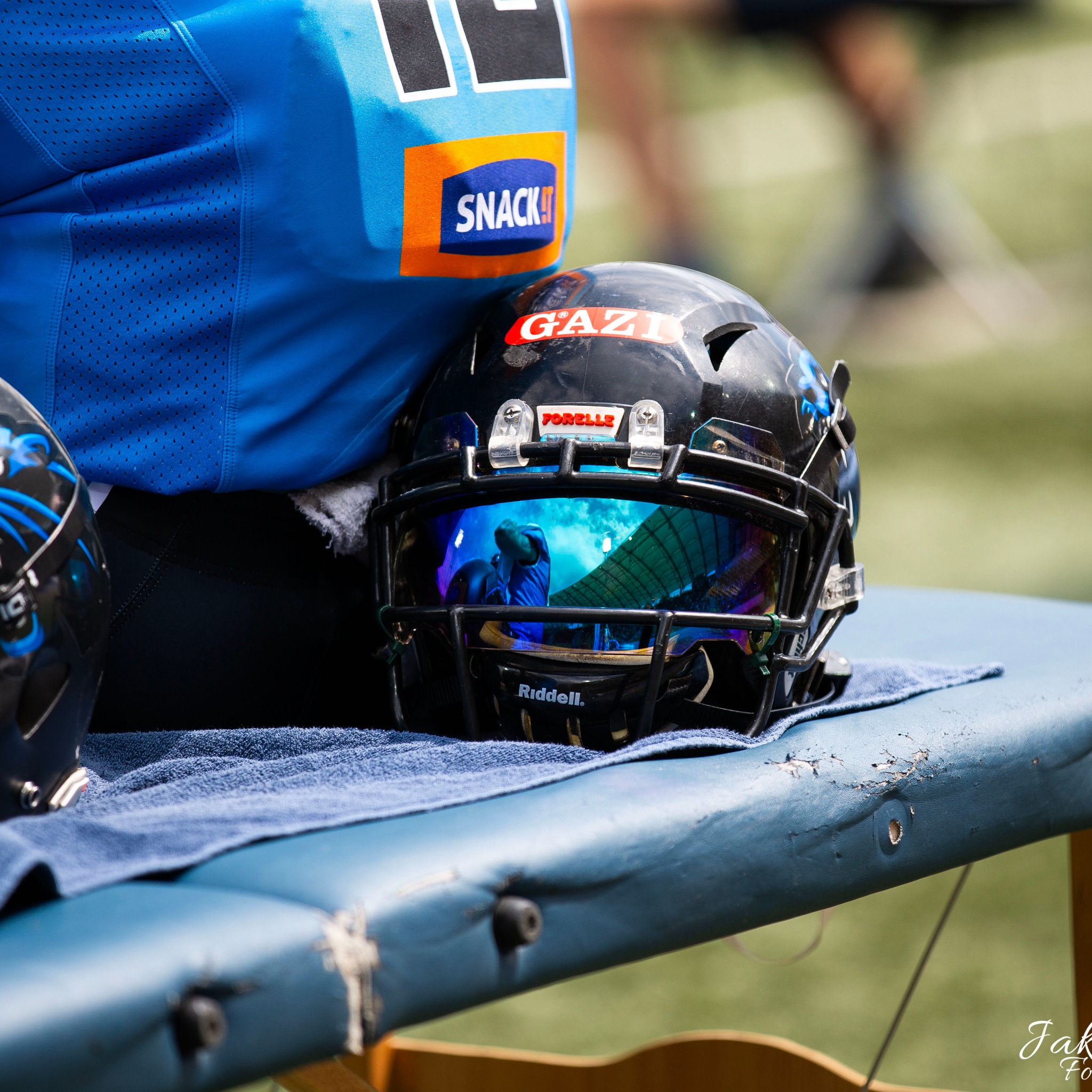 Kask futbolowy z odblaskowym wizjerem leżący na ławce obok zawodnika w niebieskiej koszulce z reklamą Snack It, widoczne odbicie stadionu w wizjerze.