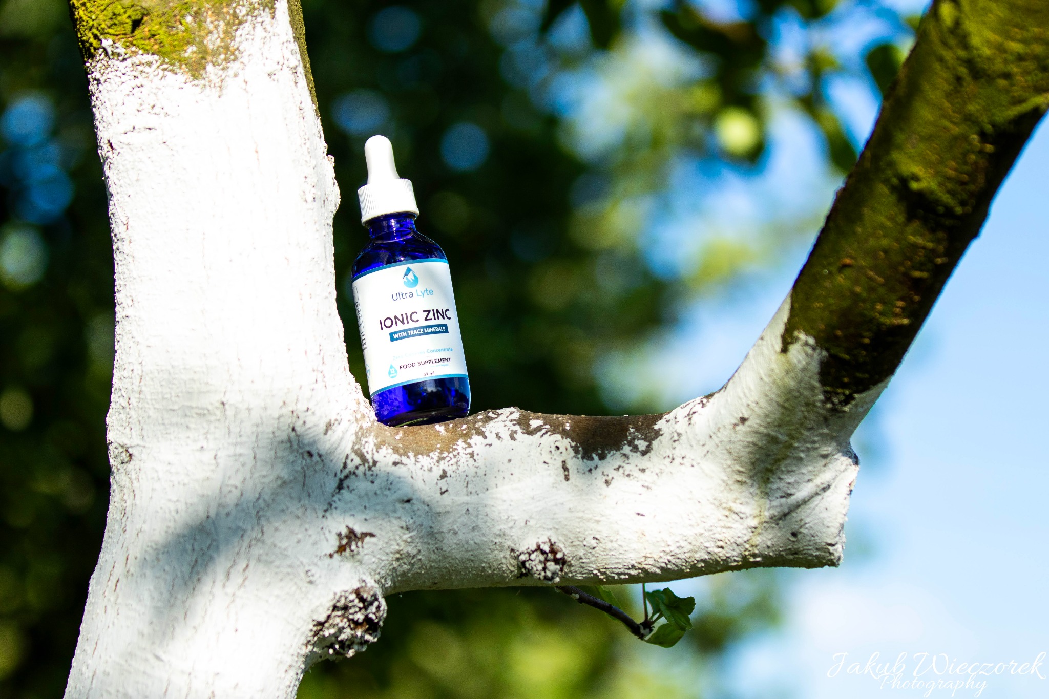 Butelka z kroplomierzem Ionic Zinc Ultra Lyte na pomalowanej na biało gałęzi drzewa, z zielonym tłem z rozmytych liści i błękitnym niebem