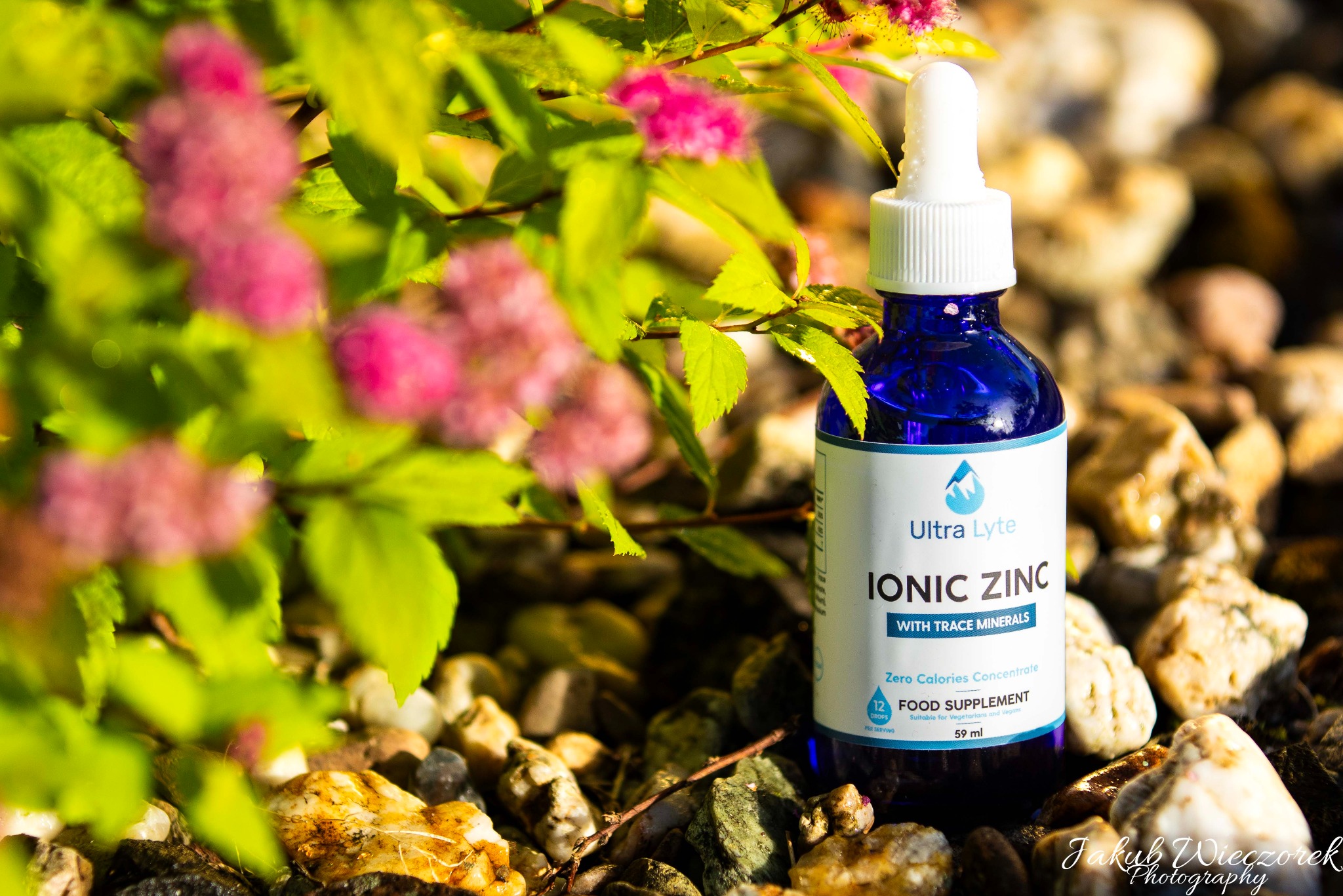 Błękitna butelka z kroplomierzem, etykieta 'Ionic Zinc', otoczona drobnymi kamieniami i kwitnącym krzewem z różowymi kwiatami.