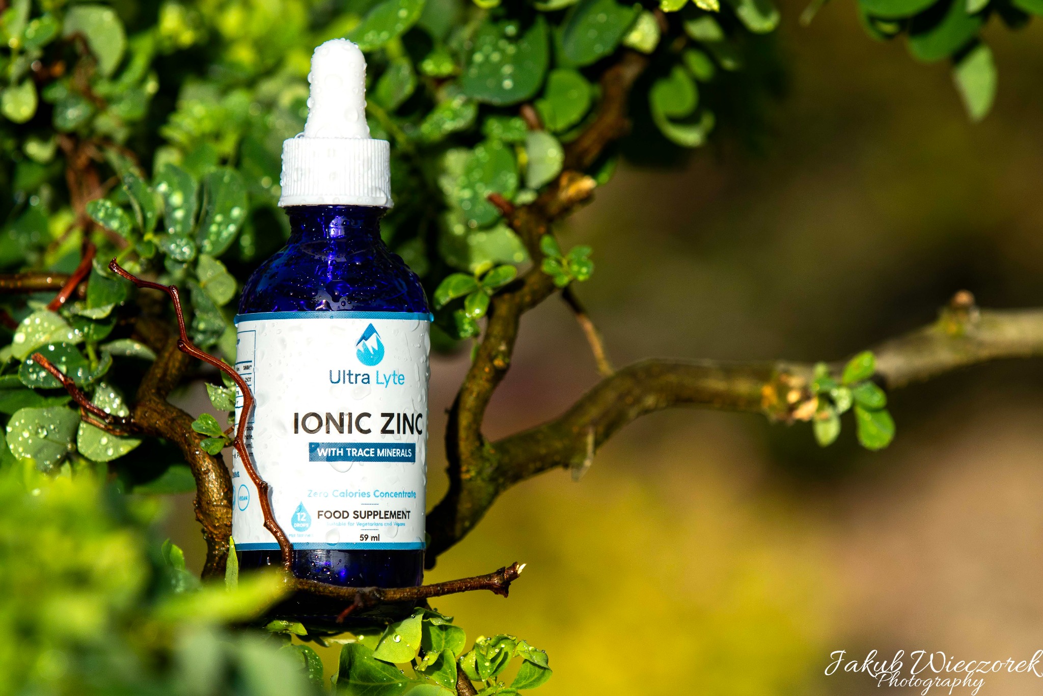 Butelka granatowego płynu Ionic Zinc Ultra Lyte z kroplomierzem, umieszczona na gałęzi zielonego krzewu z kroplami wody, na rozmytym tle.