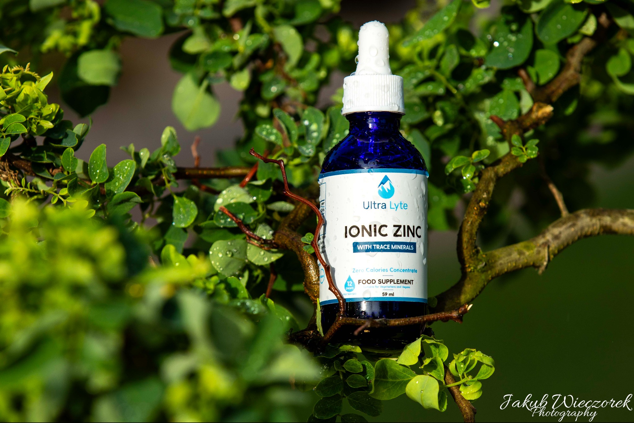 Niebieska butelka Ionic Zinc Ultra Lyte z kroplomierzem, umieszczona na tle gałązek z zielonymi liśćmi pokrytymi kroplami wody, w naturalnym oświetleniu.