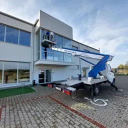 Pracownik w niebieskim ubraniu na podnośniku koszowym przy nowoczesnym budynku z dużymi oknami, widoczny numer telefonu kontaktowego na ramieniu podnośnika.