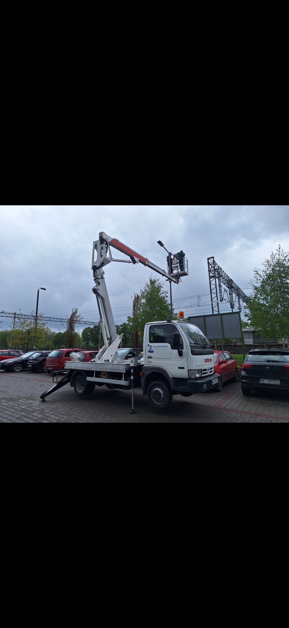 Biała ciężarówka z podnośnikiem koszowym na parkingu, wysięgnik podniesiony, w tle widoczne drzewa i konstrukcja stalowa.