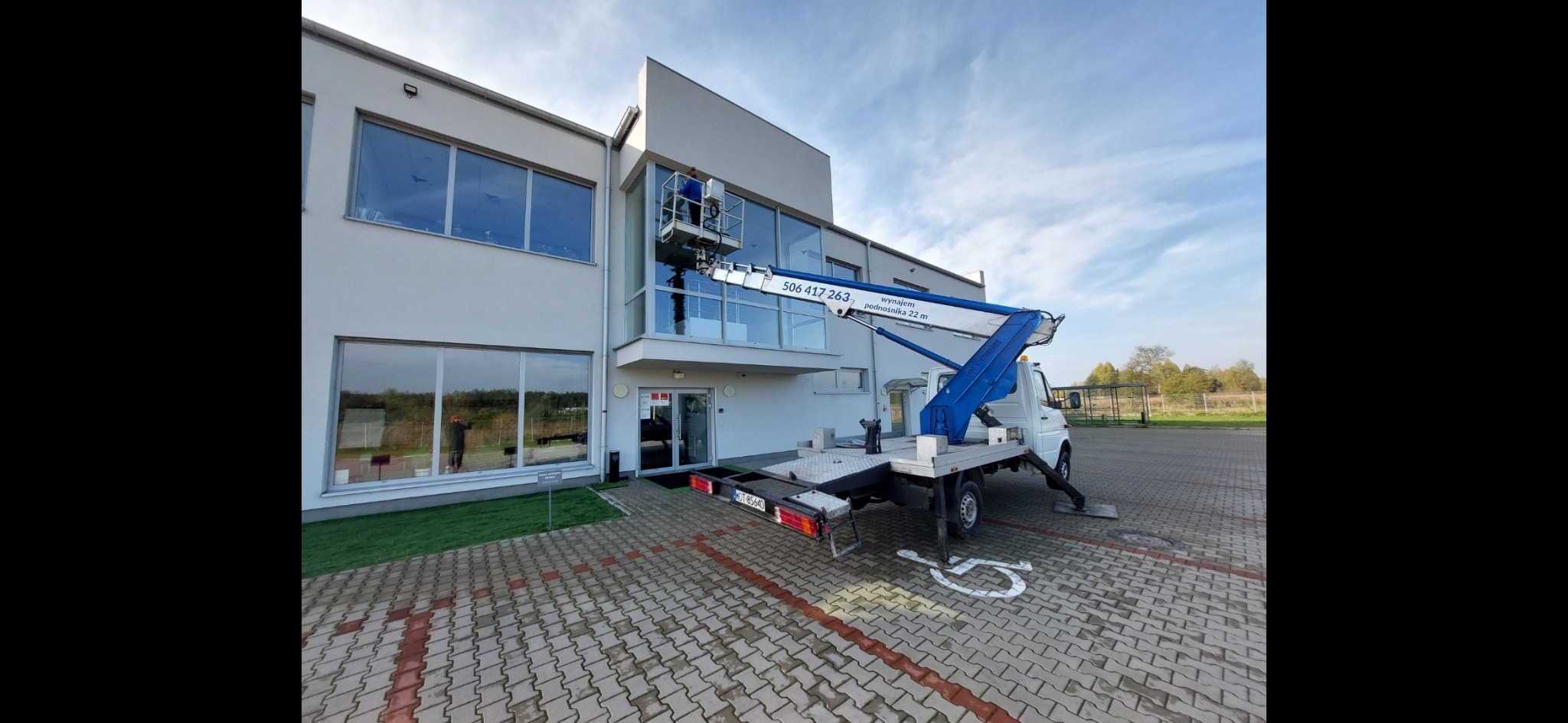 Pracownik w niebieskim ubraniu na podnośniku koszowym przy nowoczesnym budynku z dużymi oknami, widoczny numer telefonu kontaktowego na ramieniu podnośnika.